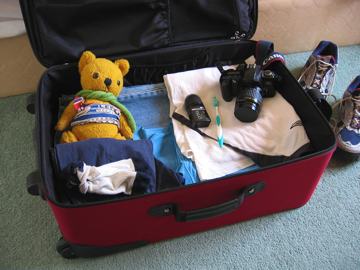 Photo of a suitcase being packed for holiday. It contains clothes, camera gear and a soft toy.