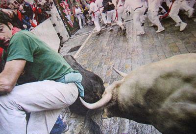 Photo of a tourist in Spain attending the annual running of the bulls. In the background are many people running and looking behind them. In the foreground the man’s left knee is bent right up next to his elbow. He is looking in horror behind him at the tip of the horn of a muscular bull which is currently millimeters away from ripping him a second arsehole.