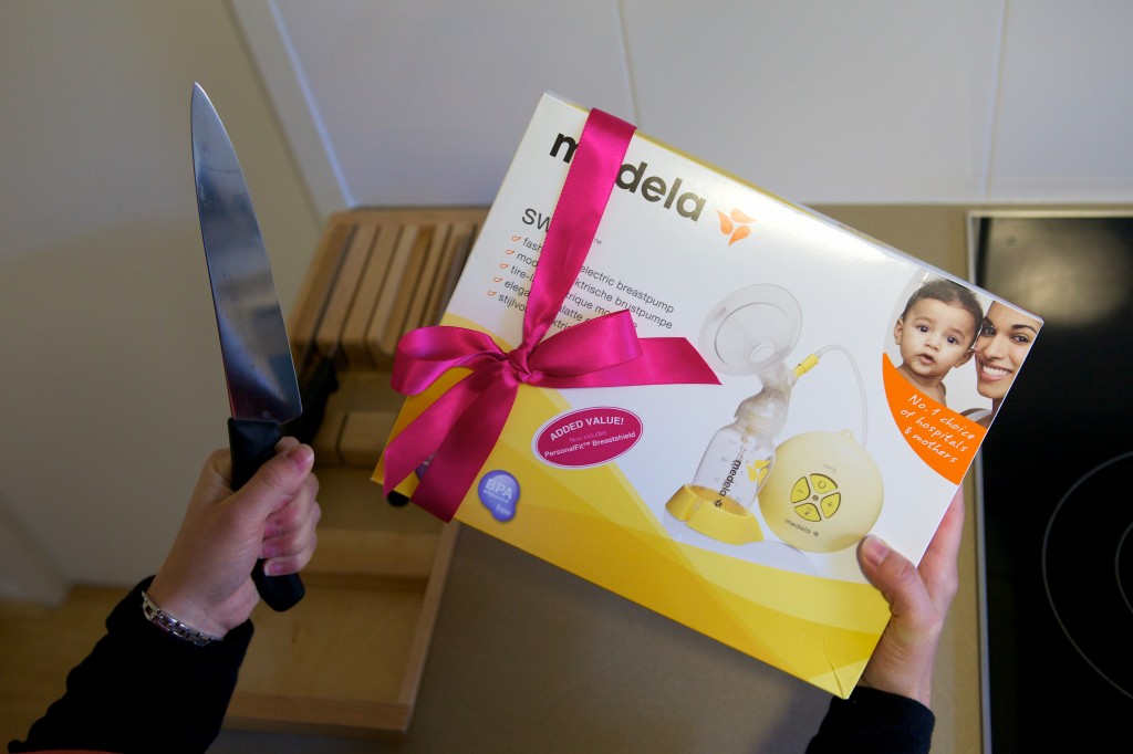 Photo of a breast pump still in the box with a red ribbon wrapped around it. It is being held in the recipient’s right hand while their left hand holds a large kitchen knife.