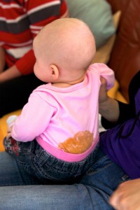 Photo of a young, sitting baby taken from behind. There is a large brown stain that starts at the baby’s bottom and stretches halfway up their back.