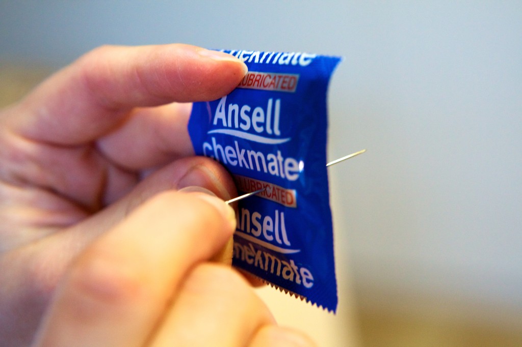 Close up photo of a female’s fingers holding a wrapped condom while pushing a pin through the centre of it.