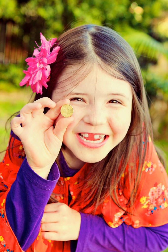 Close up photo of a smiling young girl holding a 2 dollar coin up next to the space where her tooth used to be.