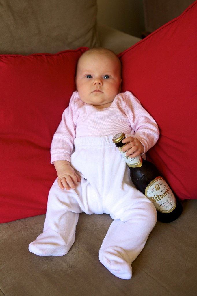 Photo of our 2 month old daughter sitting on the lounge with an unopened bottle of German beer propped between her legs.