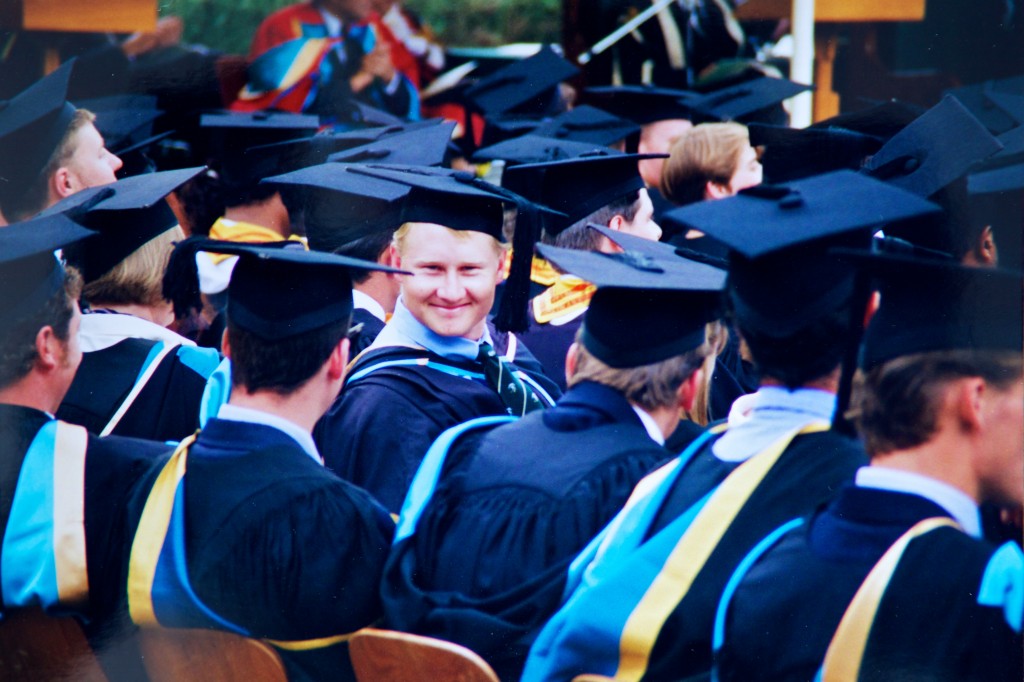 Photo of a graduate wearing cap and gown in a crowd of others. All the graduates are looking away from the camera except for the guy in the middle who is looking straight at the camera and is obviously pretty happy with himself.