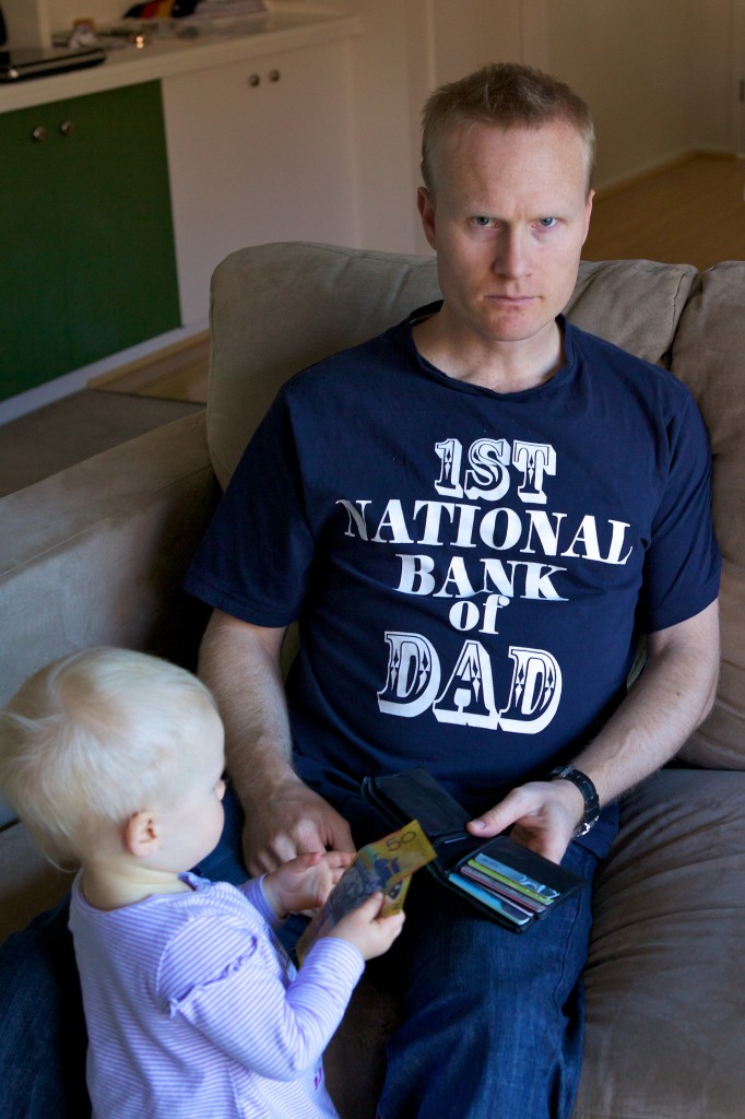 Photo of myself holding an open wallet handing our daughter a fifty dollar note. I have a look of annoyance on my face and am wearing a shirt that reads: First National Bank of Dad. 