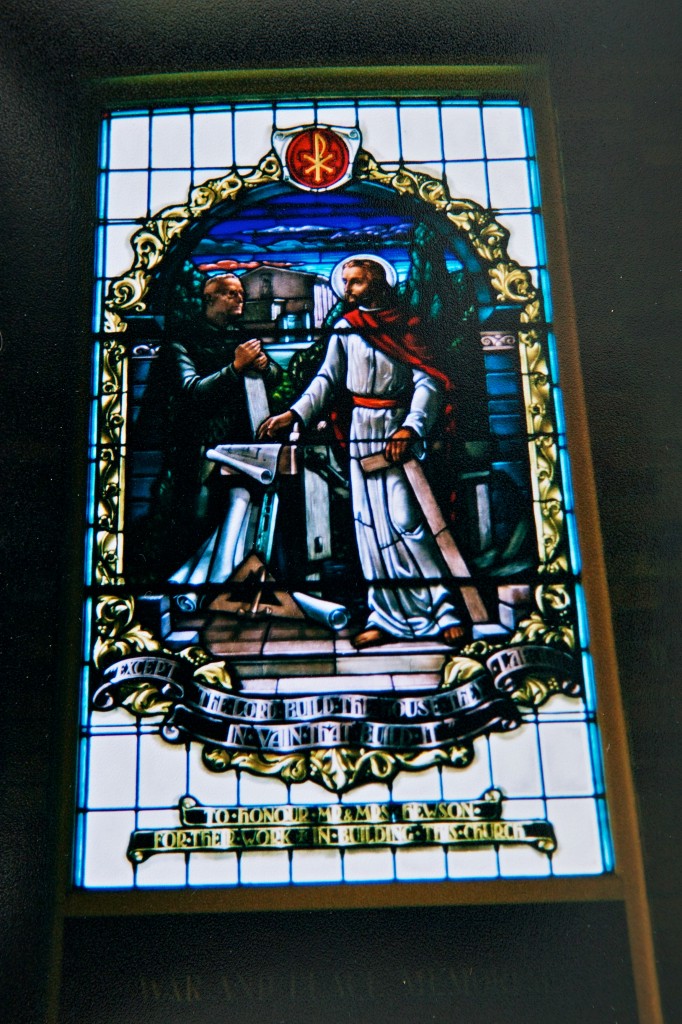 Photo of a stained glass window showing two men, one kneeling, one standing, and a table between them with what looks like building plans.