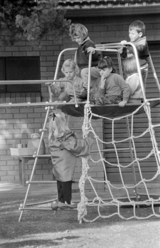 A black and white photo of a bunch of children playing on equipment in the grounds of a pre-school.