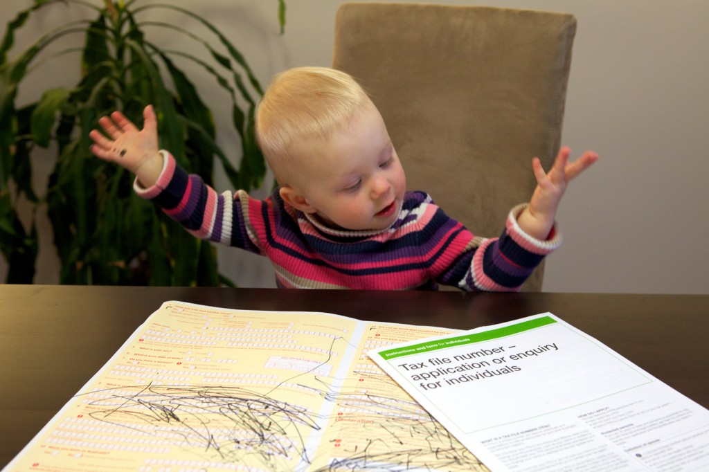 A toddler with a bemused look on her face sitting in front of a Tax File Number application form with pen scribbled randomly all over it.