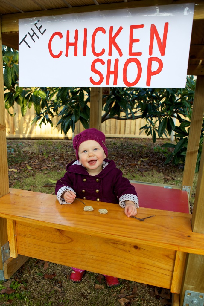 Photo showing our daughter on her play equipment in our backyard. She is standing behind a very small counter, above which is a sign that reads “The chicken shop”. On the counter are two small rocks and a stick. She is looking towards the camera with a big smile.